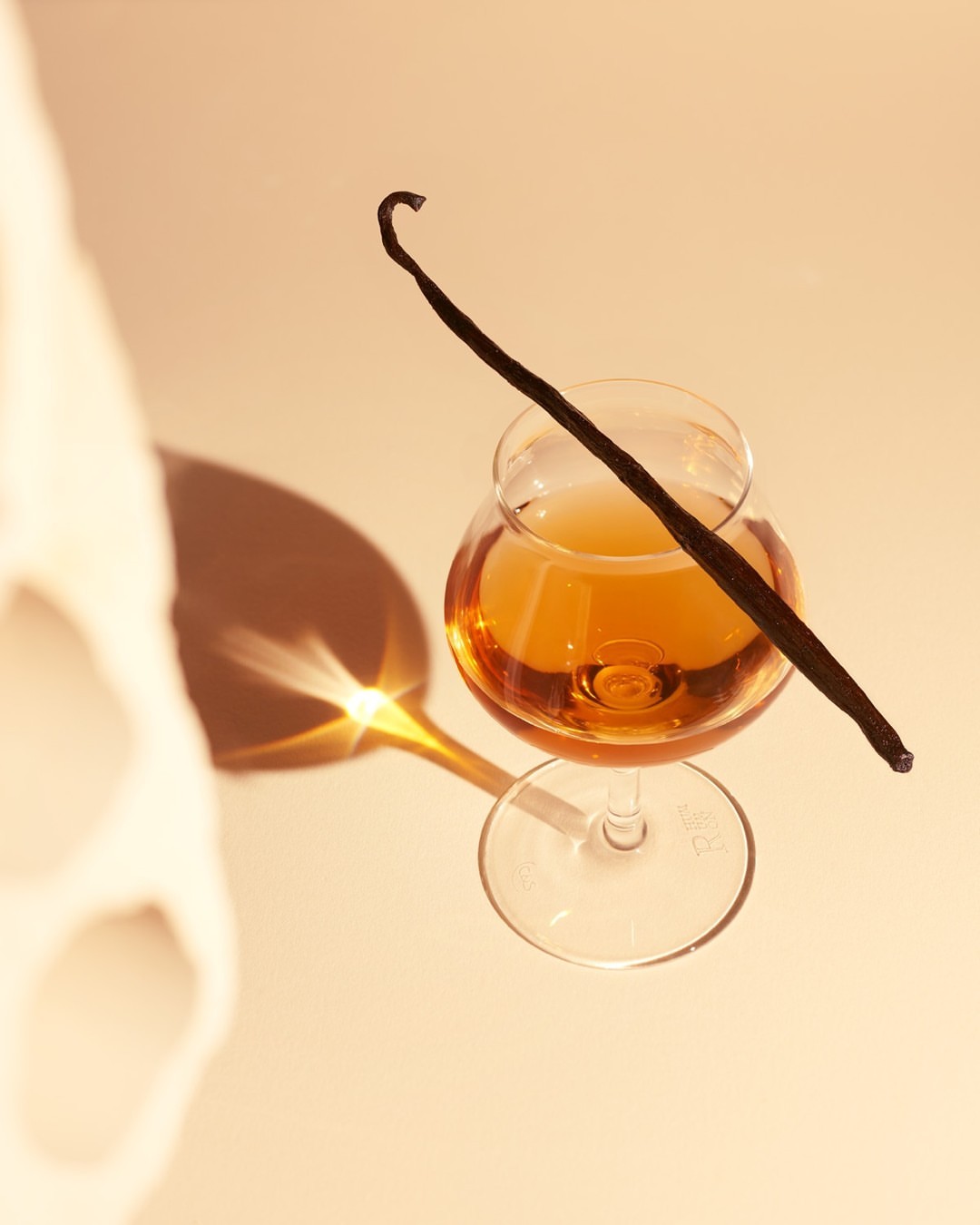 Close-up studio shot of a clear glass filled with an amber liquid and garnished with a vanilla bean on a peach-colored surface.