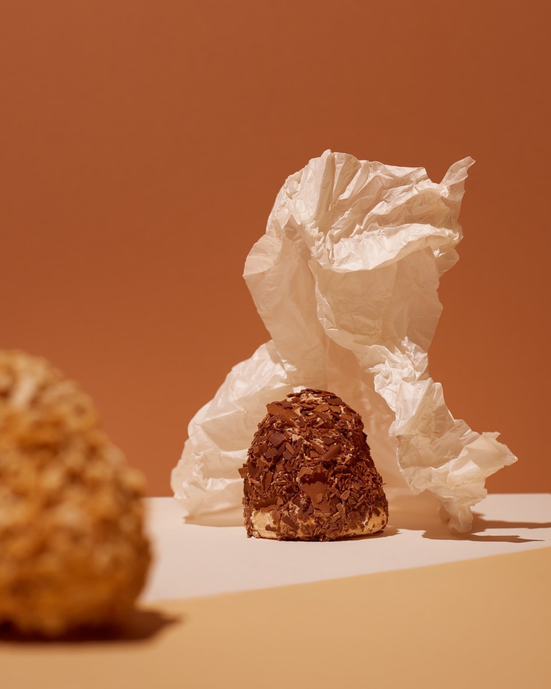 Still life of a chocolate-covered cake on a white surface, next to another cake, against a warm brown background with white crumpled paper.