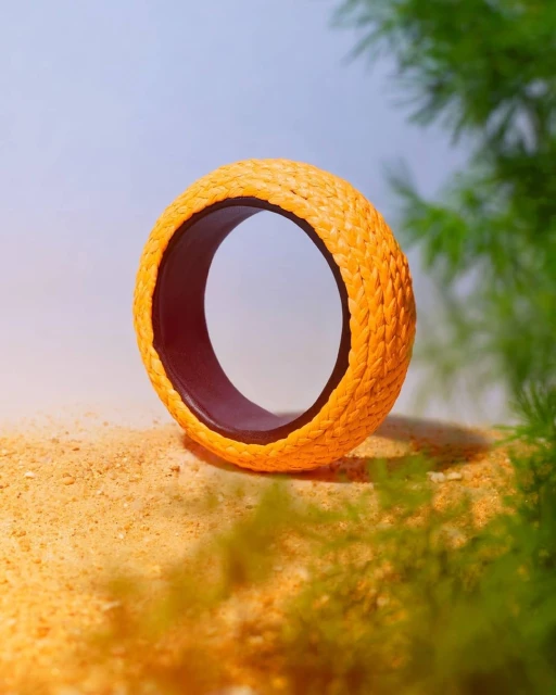 Braided orange bangle on a sand-like surface, soft green foliage in the background, lit by a soft, diffused light source.