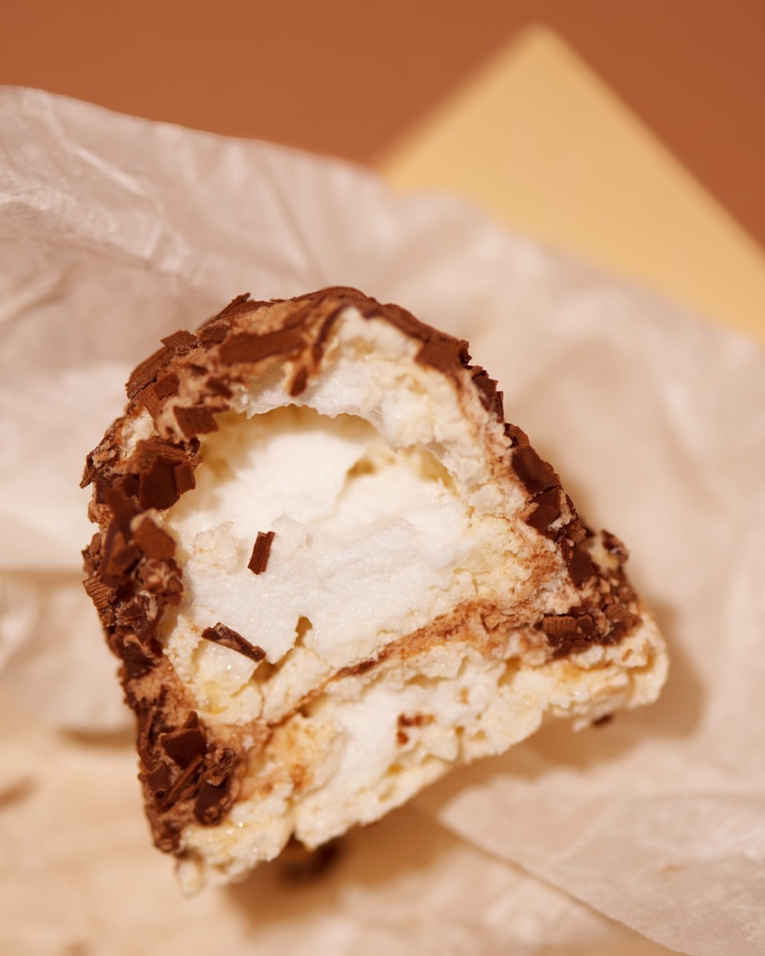 A close-up of a chocolate dessert with a white creamy filling on white parchment paper against a tan and cream background.