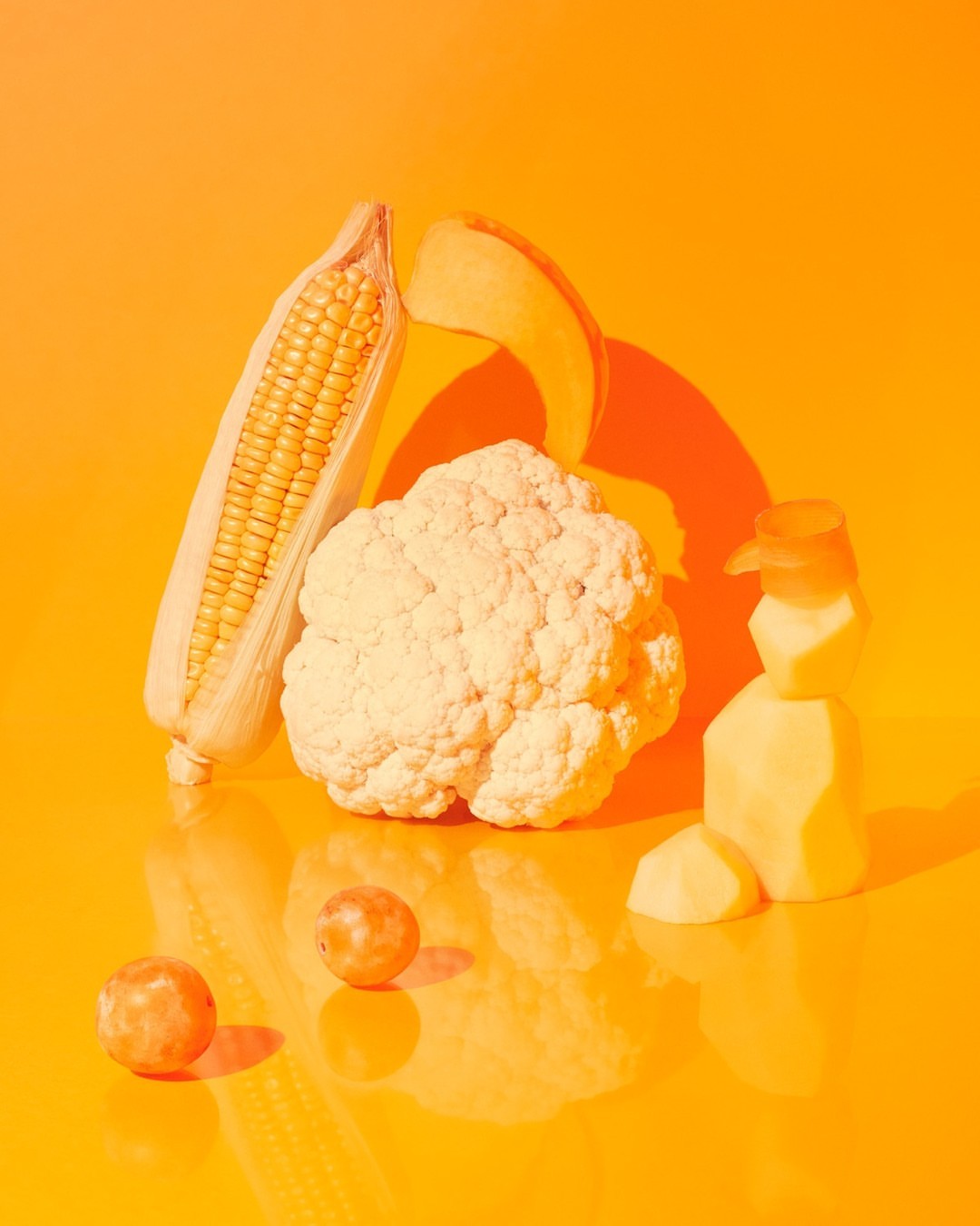 Studio shot of corn on the cob, cauliflower, grapes, potato, and pumpkin slice on reflective orange surface and backdrop.