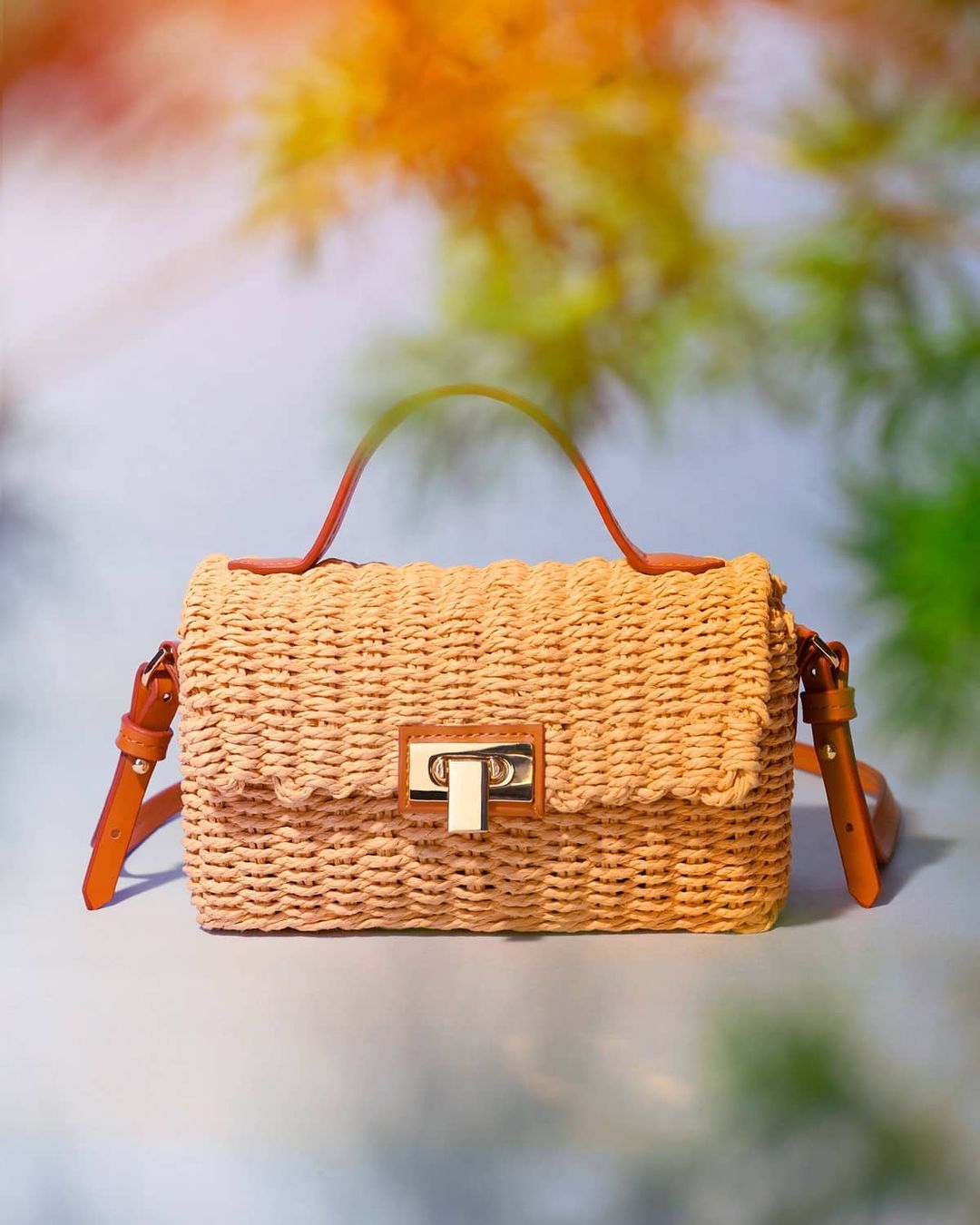 Wicker purse with leather straps, presented against a blurred backdrop of trees and sky, showcasing the bag's design and craftsmanship.