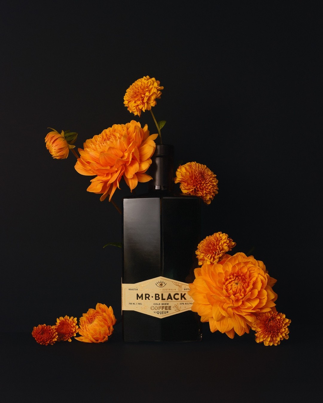 Black bottle of Mr. Black Cold Brew Coffee Liqueur surrounded by orange dahlia flowers against a dark background in studio shot.