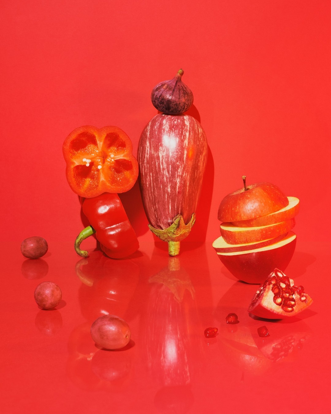 Still life studio shot on red: eggplant topped with a fig, sliced red bell pepper, sliced apple, pomegranate section, red grapes.