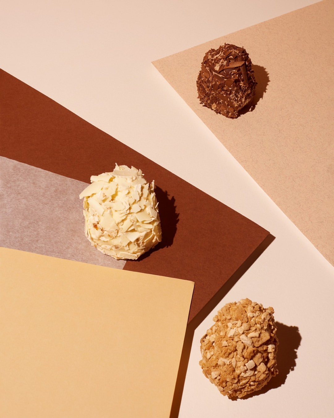 Studio shot of three chocolate truffles covered in nuts and white chocolate flakes on a tan and brown geometric background.