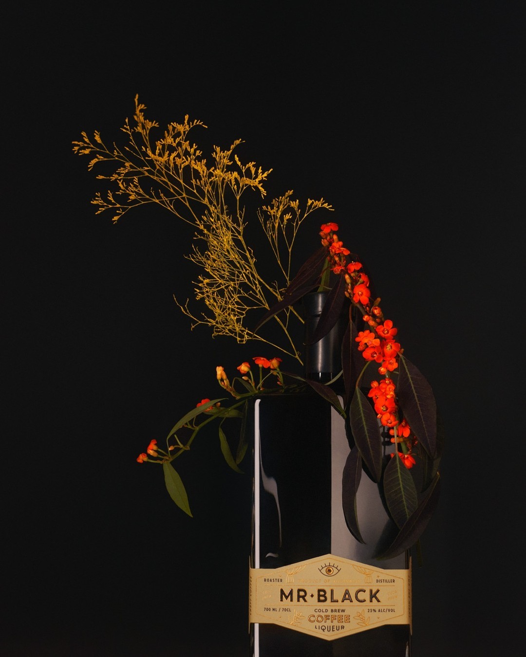 Still life photograph of a bottle of Mr. Black coffee liqueur adorned with orange flowers and golden foliage against a dark backdrop.