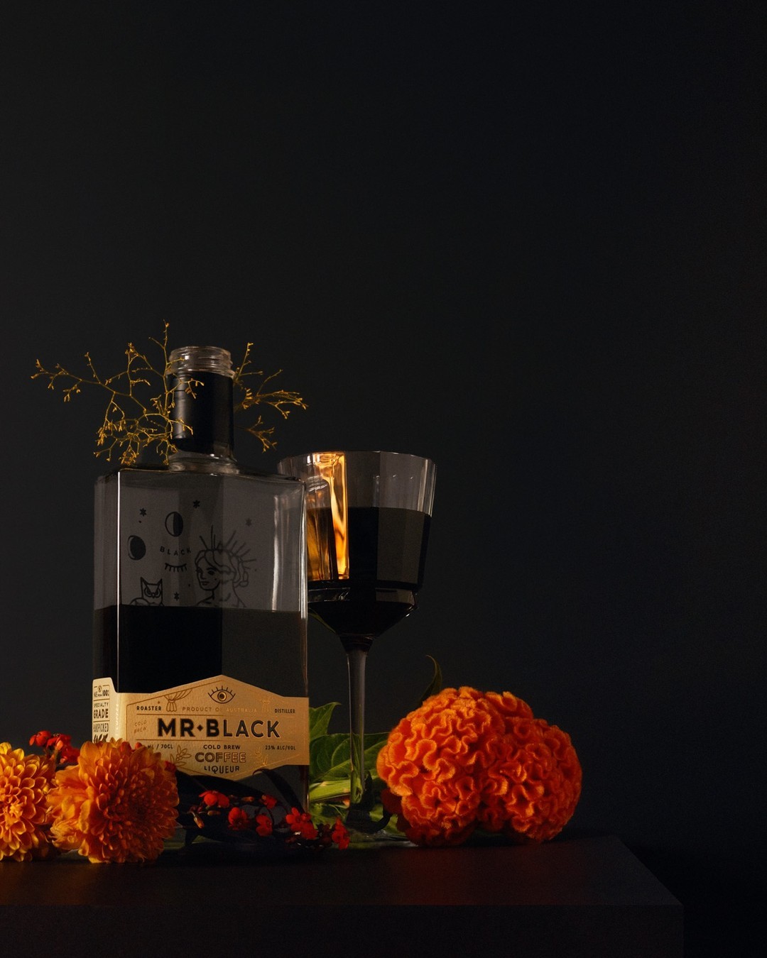 Still life of Mr. Black liquor bottle and glass with dark liquid, decorated with bright orange flowers on a dark surface.
