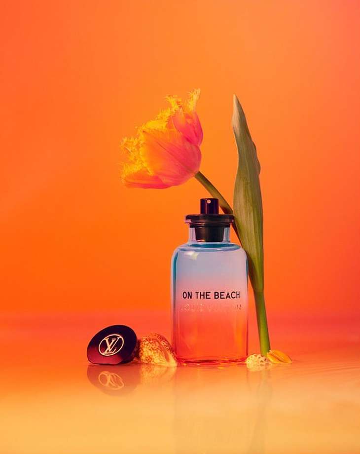 Still life of Louis Vuitton On The Beach perfume, tulip flower, and LV logo compact against a gradient orange background in studio.