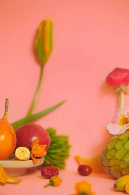 Still life food photography featuring tropical fruits like mango, cherimoya, and passion fruit arranged with flowers on a pink surface.