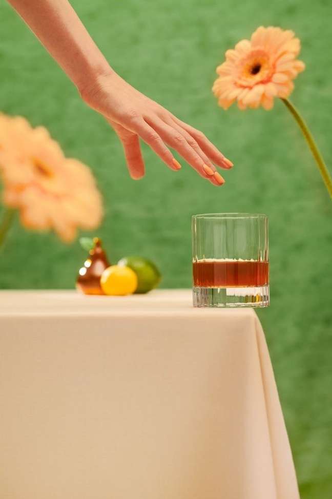 Woman's hand reaches toward a glass of brown liquid on a table with flowers, lemon, and lime against a green backdrop.