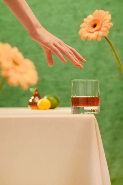 Woman's hand reaches toward a glass of brown liquid on a table with flowers, lemon, and lime against a green backdrop.