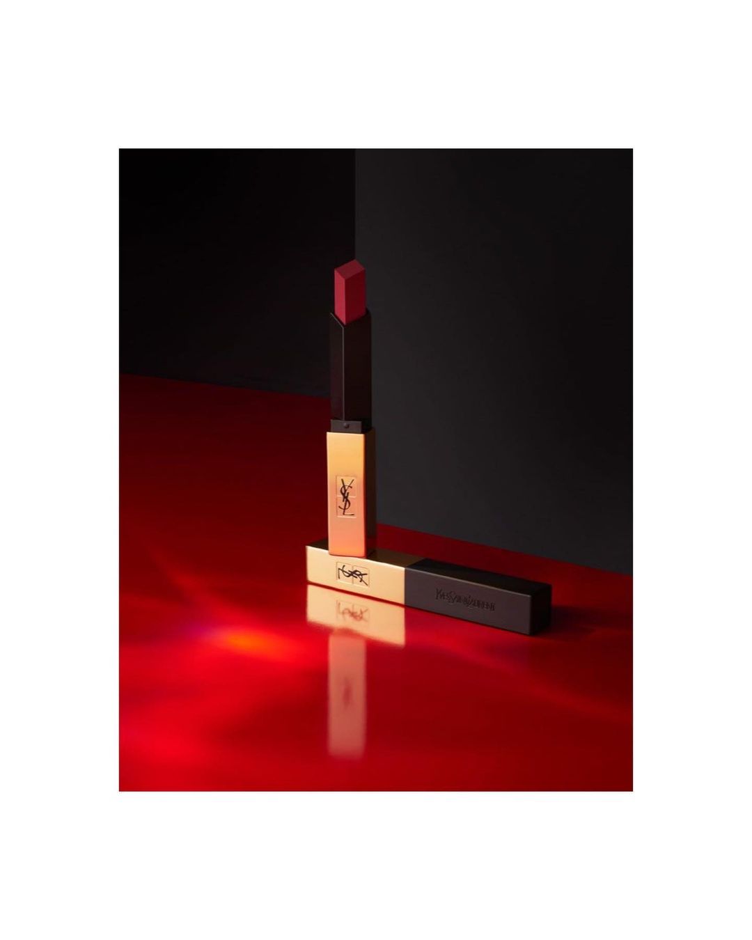 Close-up studio shot of YSL lipstick on a reflective red surface with a black background, beauty product advertisement.