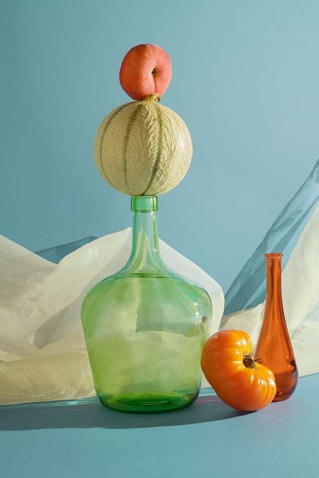 Still life photography featuring fruit: a peach atop a cantaloupe on a green glass bottle, with an orange tomato and brown bottle.
