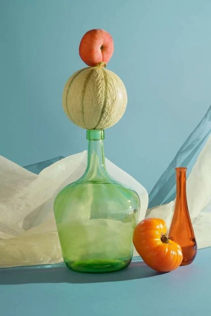 Still life photography featuring fruit: a peach atop a cantaloupe on a green glass bottle, with an orange tomato and brown bottle.