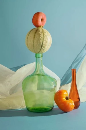 Still life photography featuring fruit: a peach atop a cantaloupe on a green glass bottle, with an orange tomato and brown bottle.