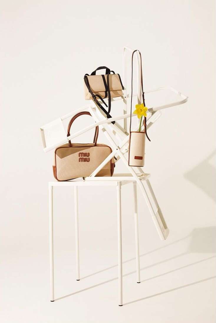 Three Miu Miu handbags in beige tones are artfully arranged on a white folding chair and metal table in a bright, neutral studio setting.