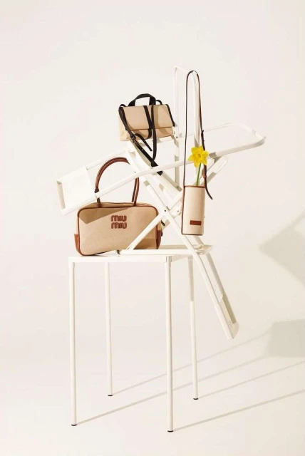 Three Miu Miu handbags in beige tones are artfully arranged on a white folding chair and metal table in a bright, neutral studio setting.
