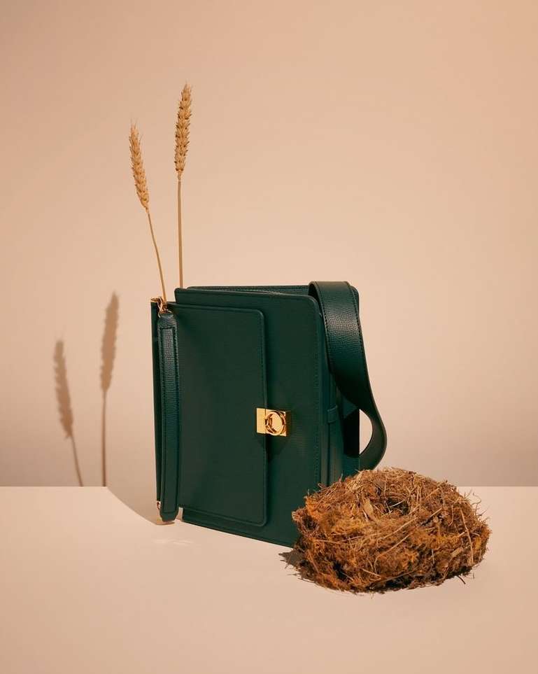 Stylish forest green satchel bag with golden clasp, accessorized with wheat stalks and a natural twig nest on a neutral backdrop.