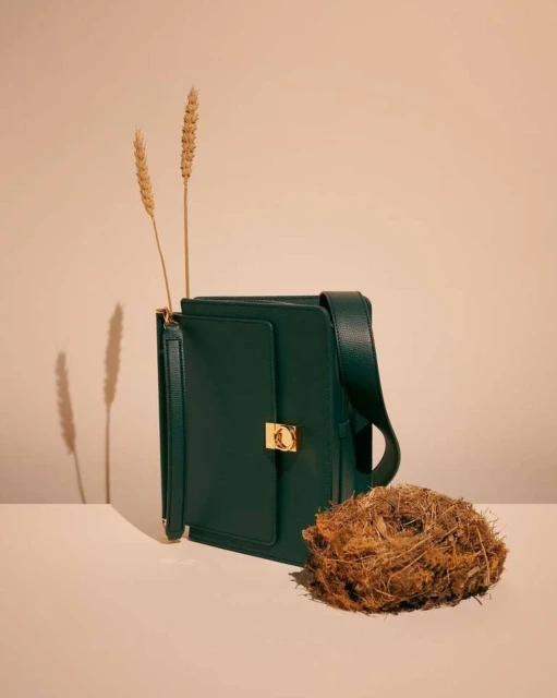 Stylish forest green satchel bag with golden clasp, accessorized with wheat stalks and a natural twig nest on a neutral backdrop.
