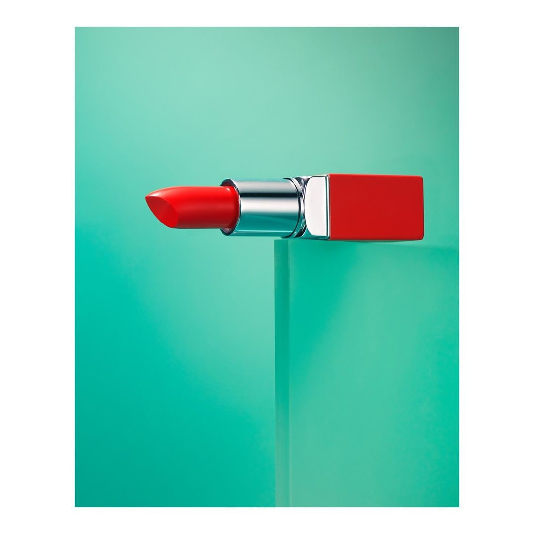 Close-up studio shot of a vibrant red lipstick with a metallic silver base on a glossy turquoise cube against a matching backdrop.