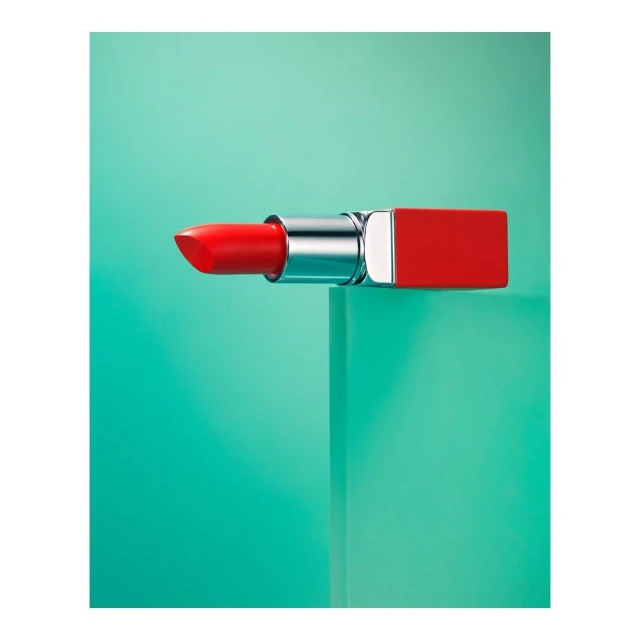 Close-up studio shot of a vibrant red lipstick with a metallic silver base on a glossy turquoise cube against a matching backdrop.