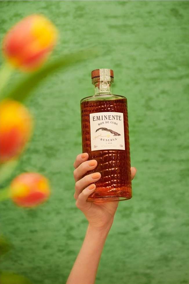 Hand holds Eminente rum bottle against green backdrop with blurred tulips. Focus is on bottle's label and design. 