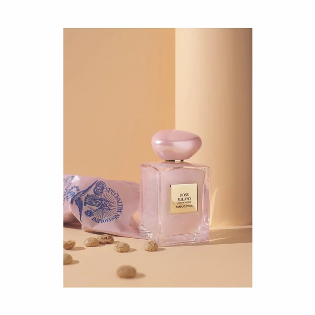 A pink perfume bottle by Armani Privé sits next to a patterned pink package and small round chocolates on a tan surface with peach backdrop.