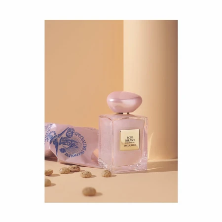 A pink perfume bottle by Armani Privé sits next to a patterned pink package and small round chocolates on a tan surface with peach backdrop.