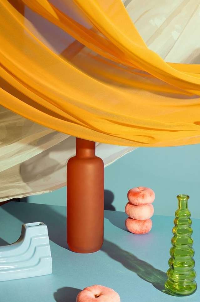 Still life photograph of a terracotta vase, stack of peaches, and blue and green glassware set against a blue background and soft lighting.
