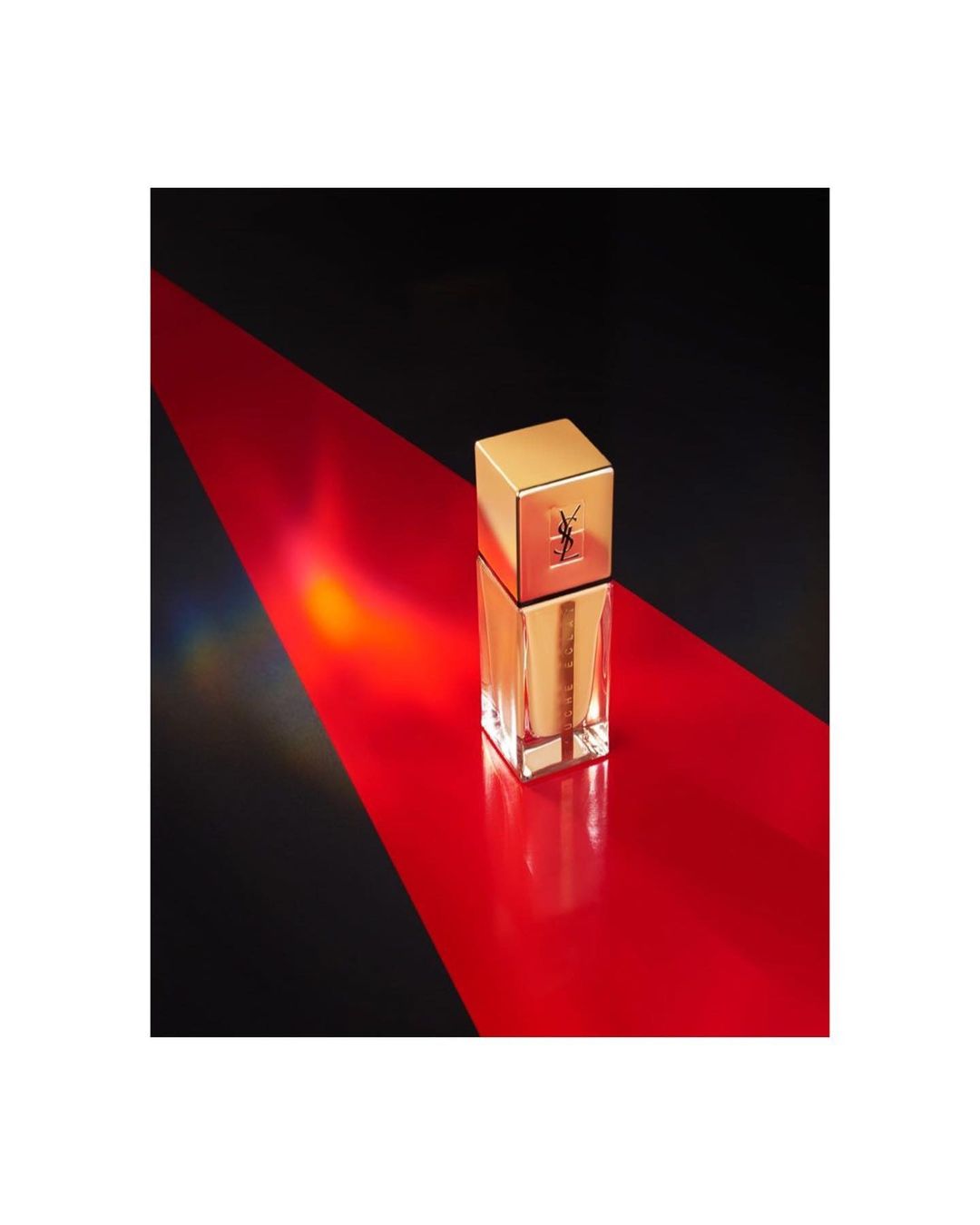 A rectangular glass cosmetic bottle with a gold cap standing upright on a glossy red surface angled against a dark background.