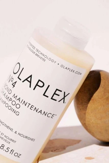 Olaplex No. 4 Bond Maintenance shampoo bottle, white label with black text, next to gold-colored stone, beauty product.