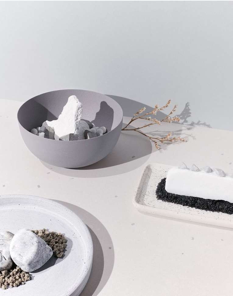 Still life featuring a gray bowl filled with abstract white shapes, a branch with small blooms, and geometric white plates with stones.