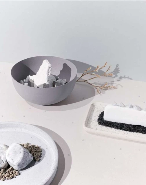Still life featuring a gray bowl filled with abstract white shapes, a branch with small blooms, and geometric white plates with stones.