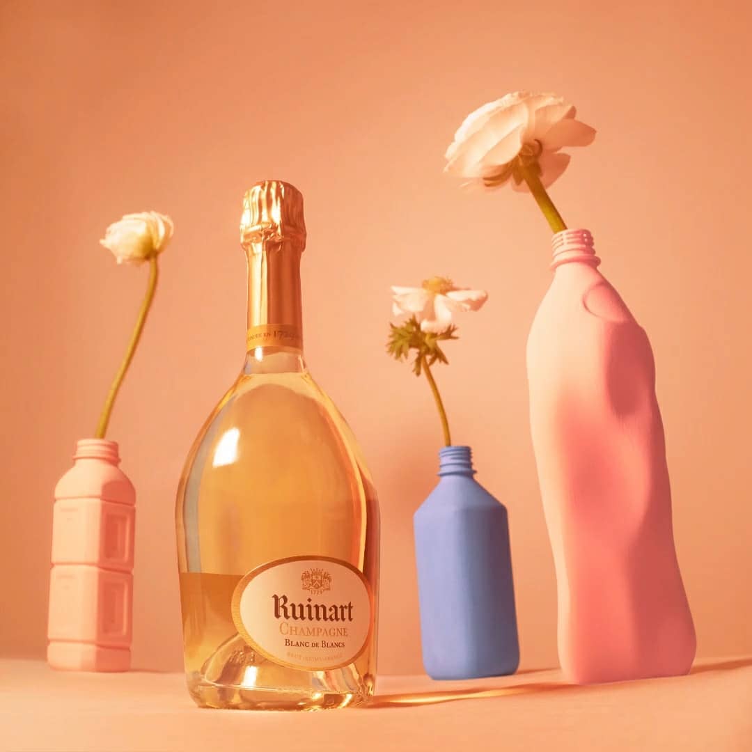Still life composition featuring a bottle of Ruinart Champagne with white flowers in colorful detergent bottles against a peach background.