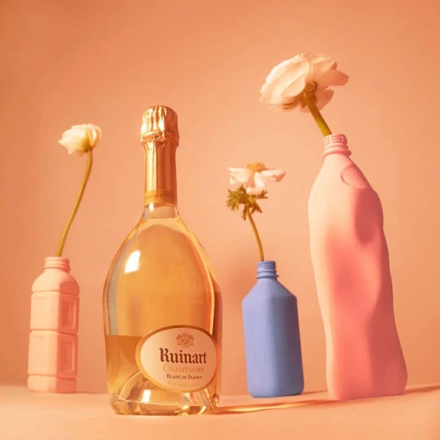 Still life composition featuring a bottle of Ruinart Champagne with white flowers in colorful detergent bottles against a peach background.