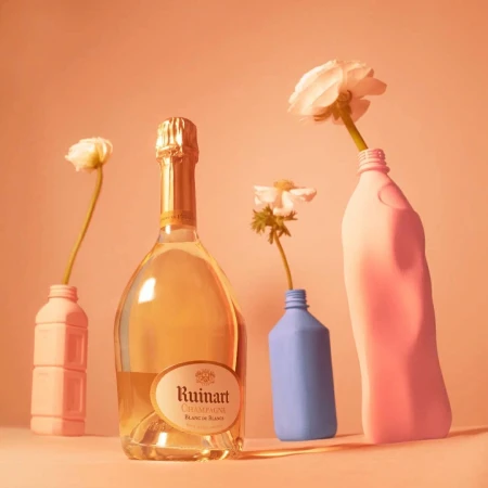 Still life composition featuring a bottle of Ruinart Champagne with white flowers in colorful detergent bottles against a peach background.