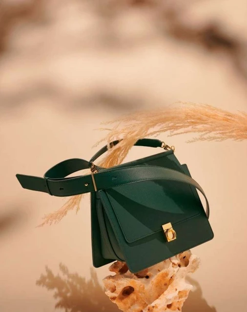 Stylish green leather handbag displayed on a textured rock with pampas grass and a neutral backdrop in a fashion editorial shot.