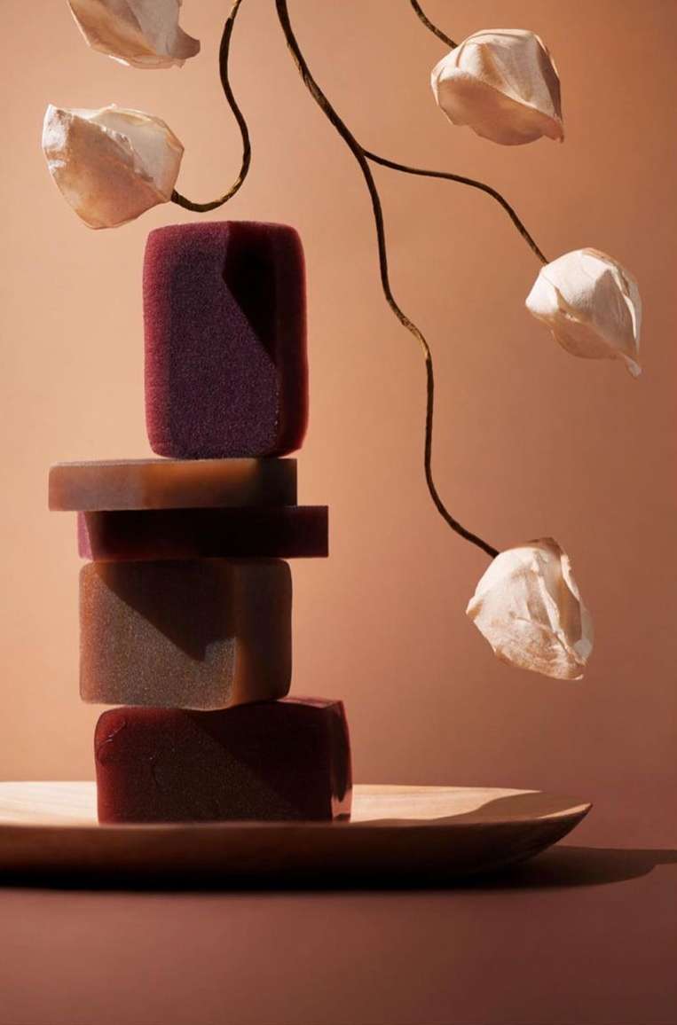 Stacked bars of soap on a wooden tray. The soap bars are different shades of dark red and brown, with some flower-shaped decorations.