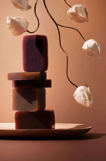 Stacked bars of soap on a wooden tray. The soap bars are different shades of dark red and brown, with some flower-shaped decorations.
