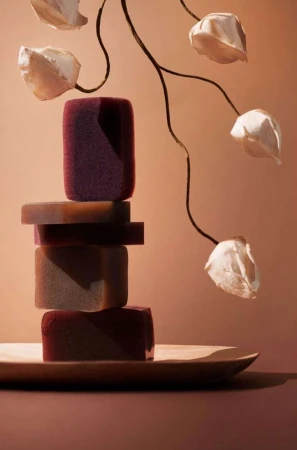 Stacked bars of soap on a wooden tray. The soap bars are different shades of dark red and brown, with some flower-shaped decorations.