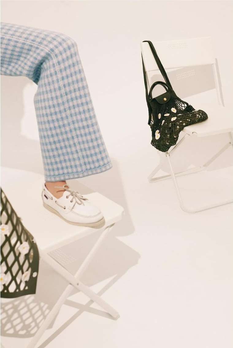 Person in blue plaid pants and white sneakers sitting on a white folding chair with a net bag and white background in a studio.