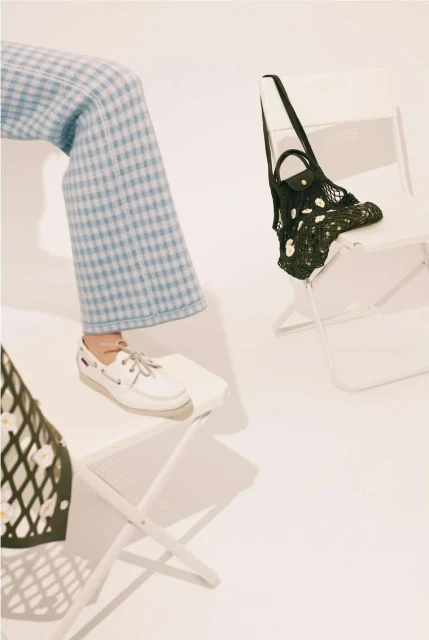 Person in blue plaid pants and white sneakers sitting on a white folding chair with a net bag and white background in a studio.