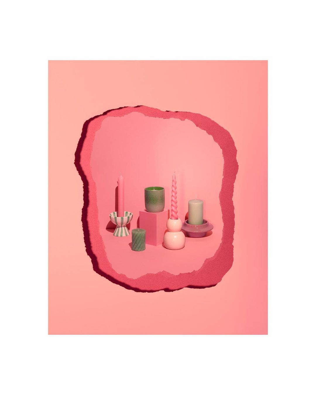 Assortment of decorative candles in various shapes and colors arranged on pink platforms against a layered pink background.