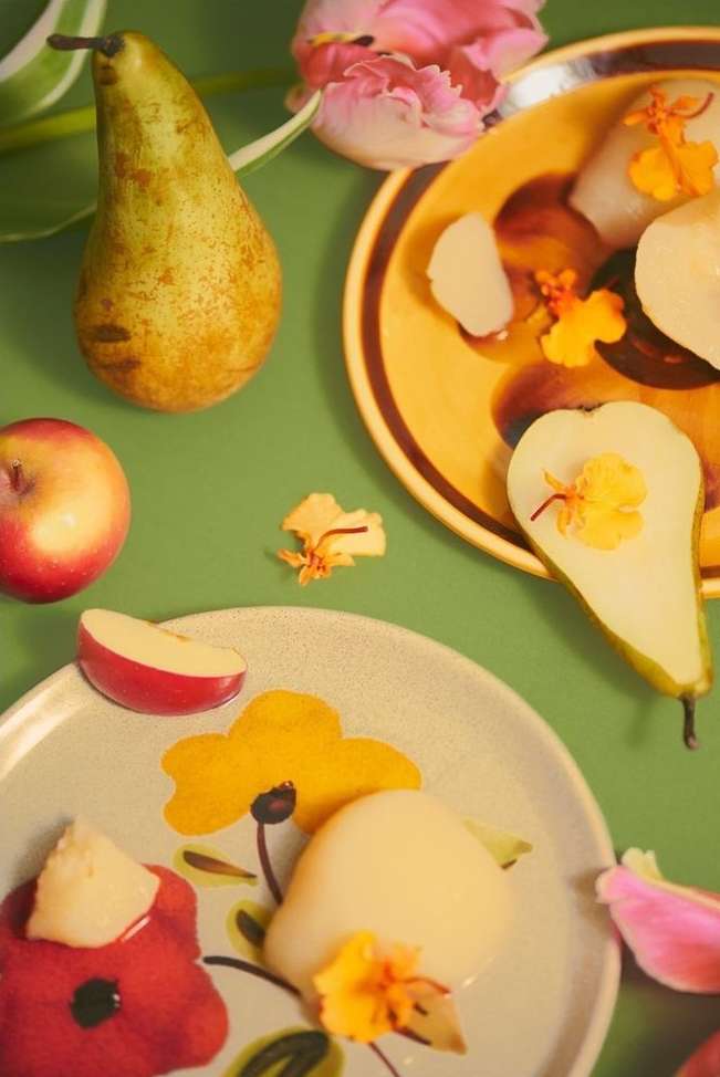 Still life of poached pears on plates with tulips and an apple on a light green surface, showcasing a food photography setup.
