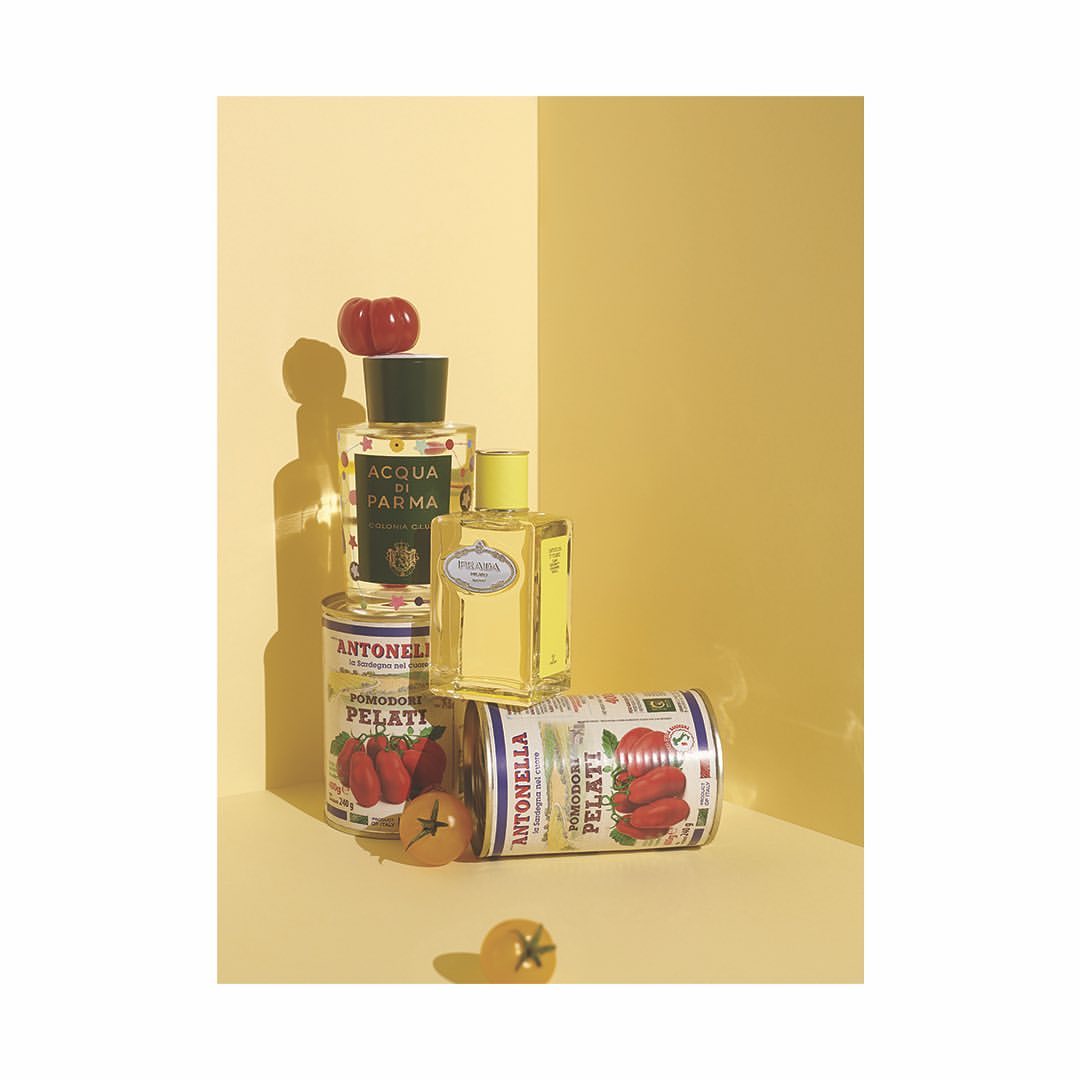 Still life shot of Acqua di Parma and Prada fragrances artfully arranged with tomato cans and fresh tomatoes against a yellow backdrop.