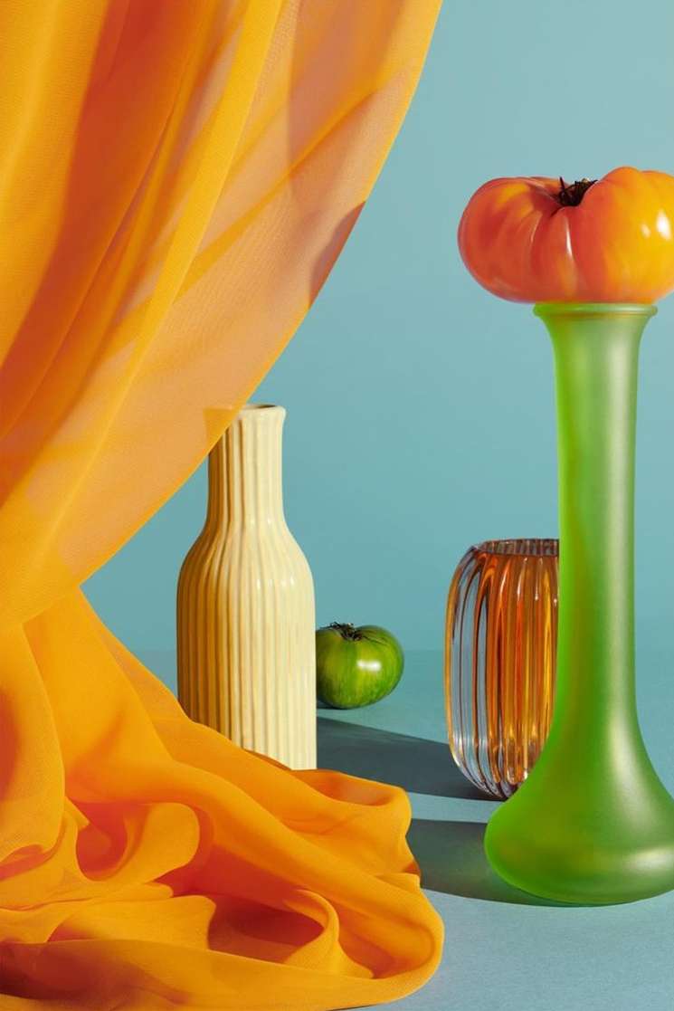Still life photograph showcasing an orange tomato atop a green vase, complemented by an off-white vase, green apple, orange vase, and orange fabric.