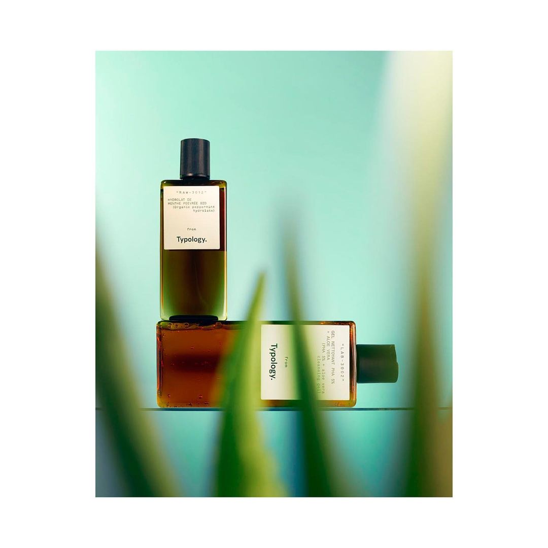 Two brown glass bottles of Typology skincare product on a light blue surface with out-of-focus green blades of grass in foreground.