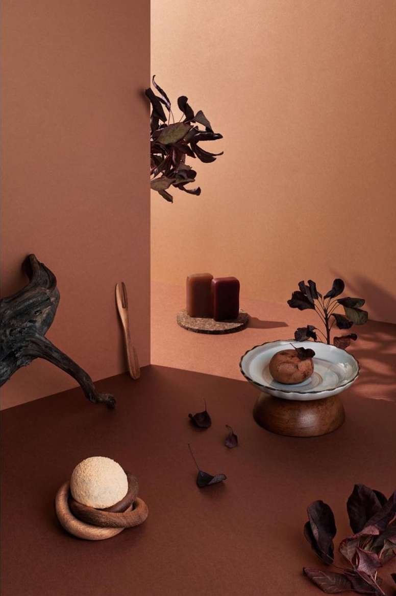 Still life composition featuring candles, decorative fruit, leaves, and driftwood arranged against a two-tone brown wall backdrop.