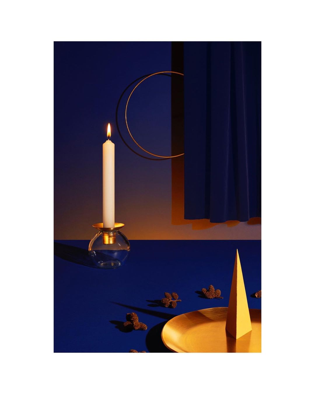 Candle in glass holder with flame, surrounded by decorative objects, on a blue surface with a dark blue curtain background.