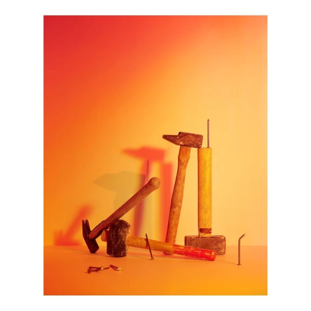 Still life composition featuring hammers, nails, and other tools against a gradient orange and yellow background in a studio setting.